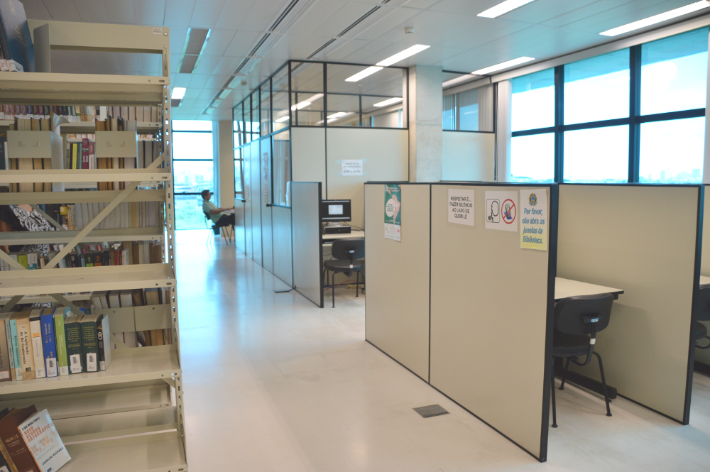 Imagem dentro da Biblioteca JFPE onde fica estante com livros e as mesas e cadeiras onde se faz pesquisas e estudos